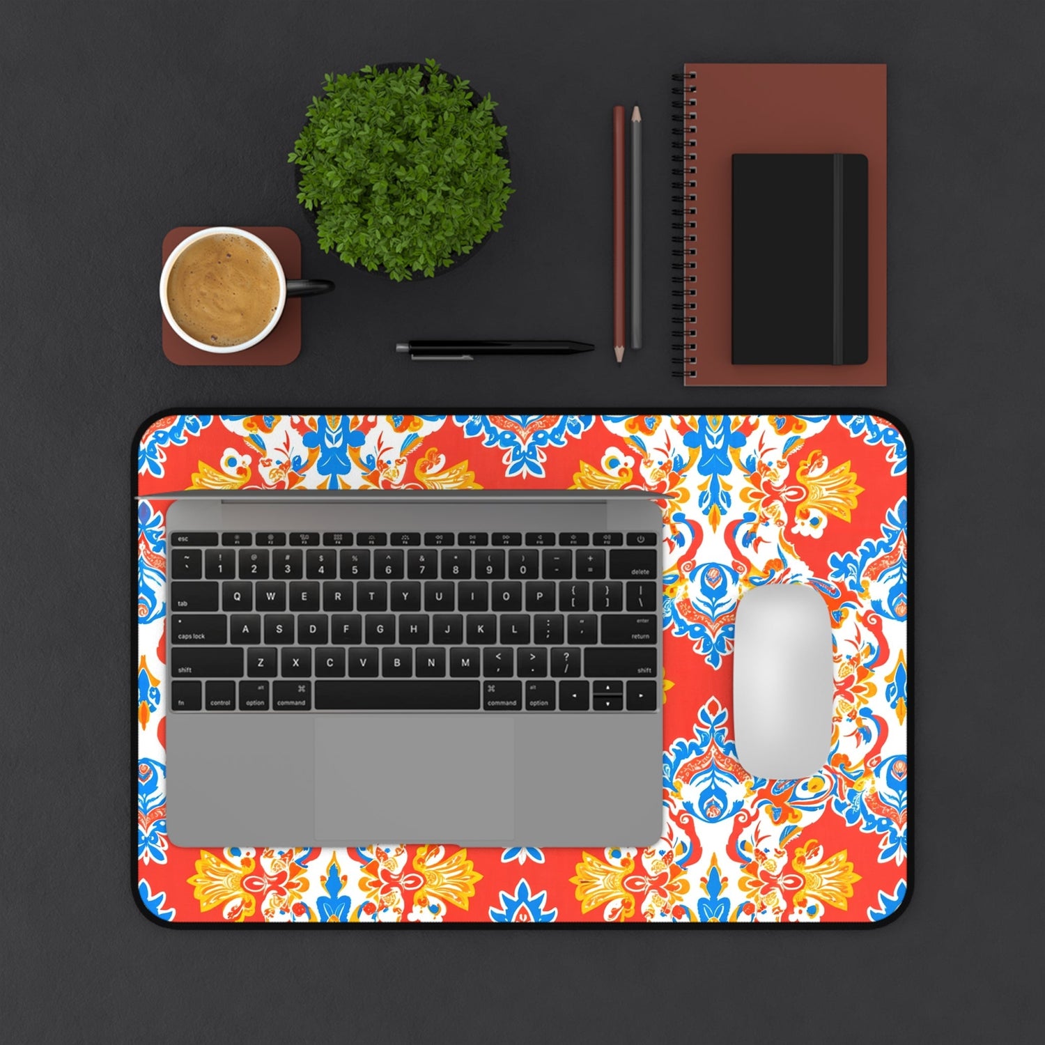 A laptop and mouse sit on a patterned mouse pad with black, white, and green designs. Nearby are a cup of coffee, a green plant, a pen, a pencil, and two stacked notebooks on a dark surface.
