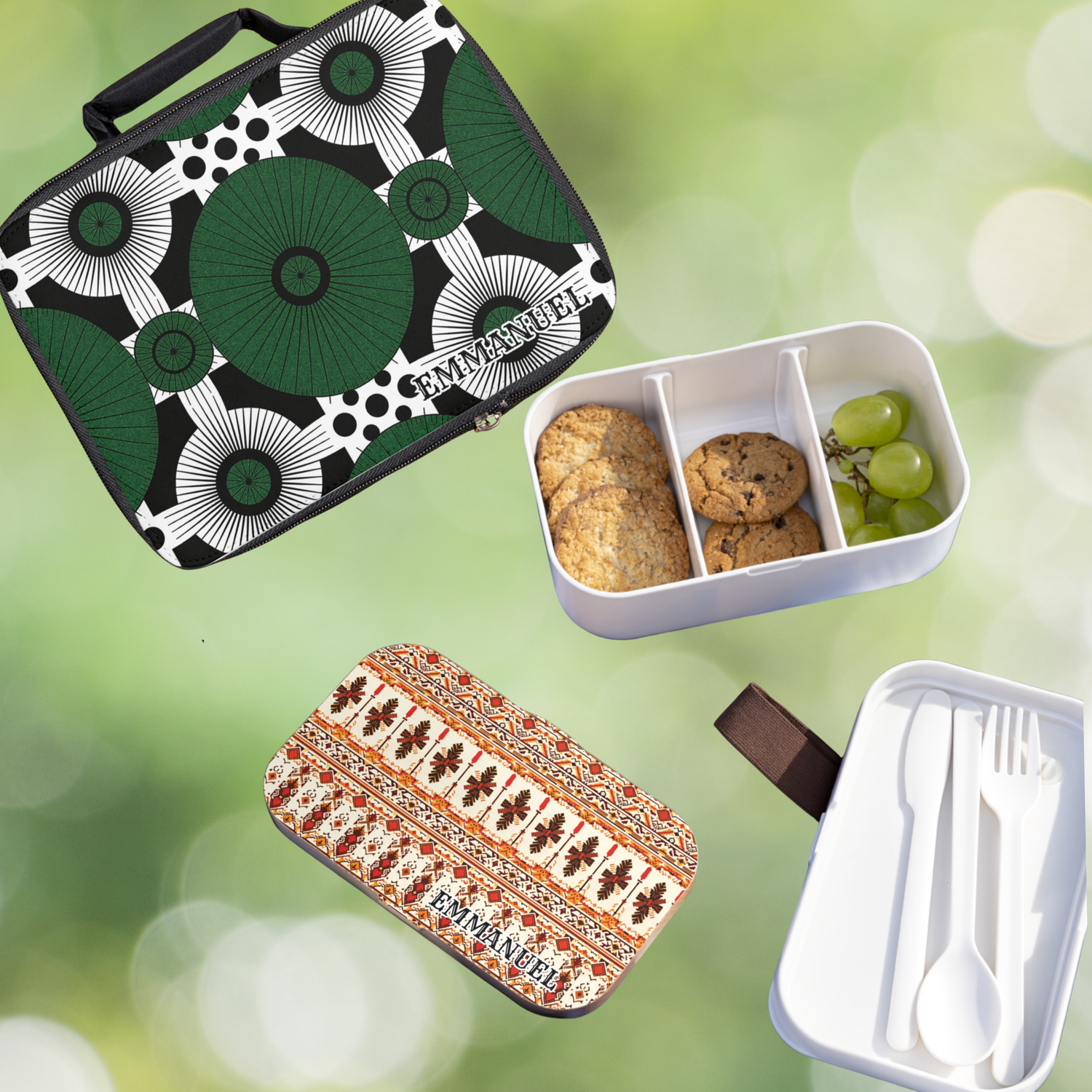 Two patterned lunchboxes labeled EMMANUEL. One is black, white, and green with cookies and grapes inside. The other is beige and orange, next to a compartment holding white cutlery. Blurred green background.