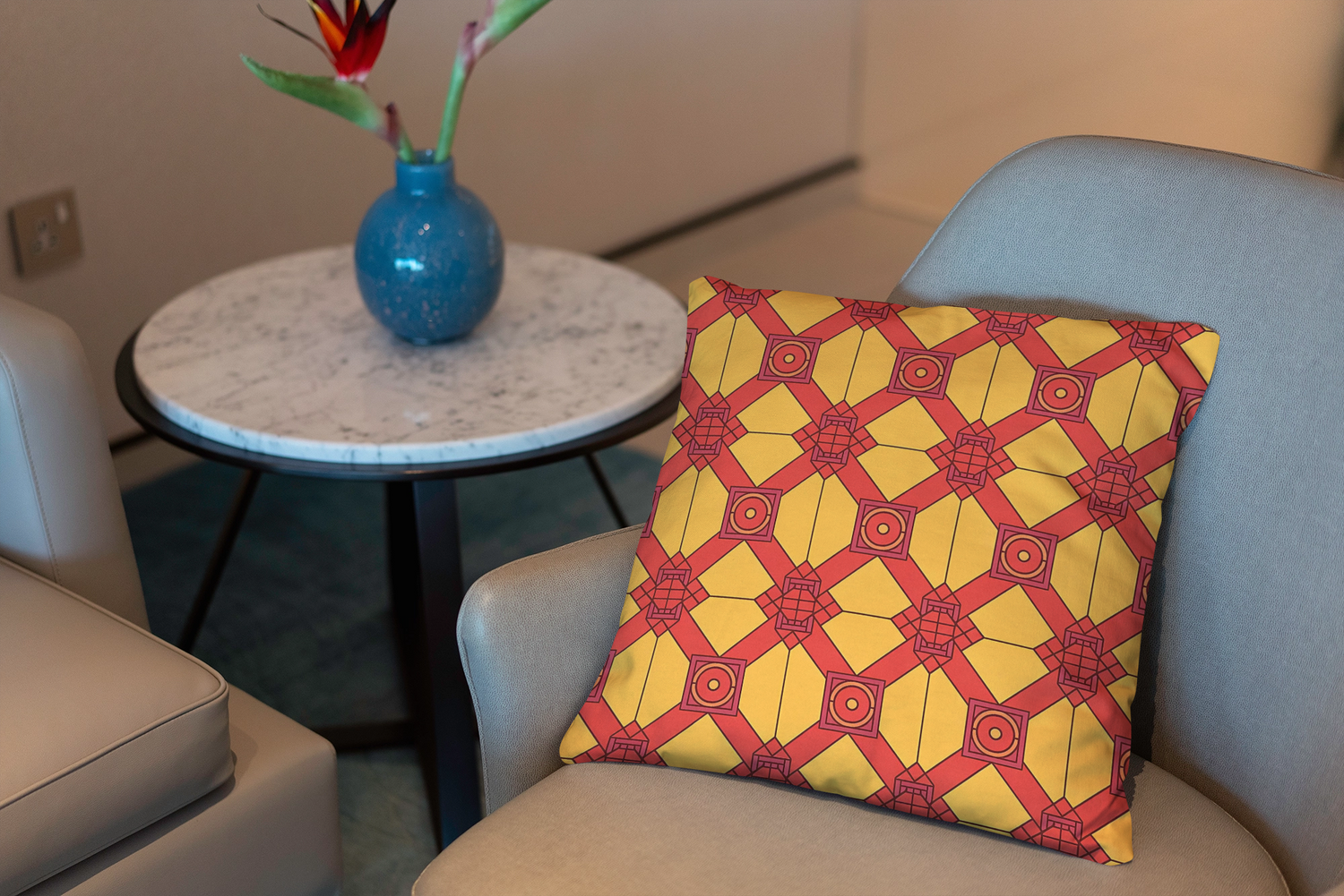 A modern beige armchair with a yellow and red geometric-patterned pillow sits by a round white marble table holding a blue vase with a bird of paradise flower.