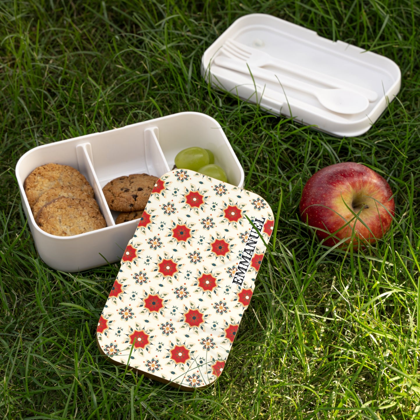 A floral-patterned, BPA-free lunchbox with three compartments—two with cookies, one with green grapes—sits on grass. The matching lid and a white utensil set are nearby. An apple rests beside this inviting picnic scene.