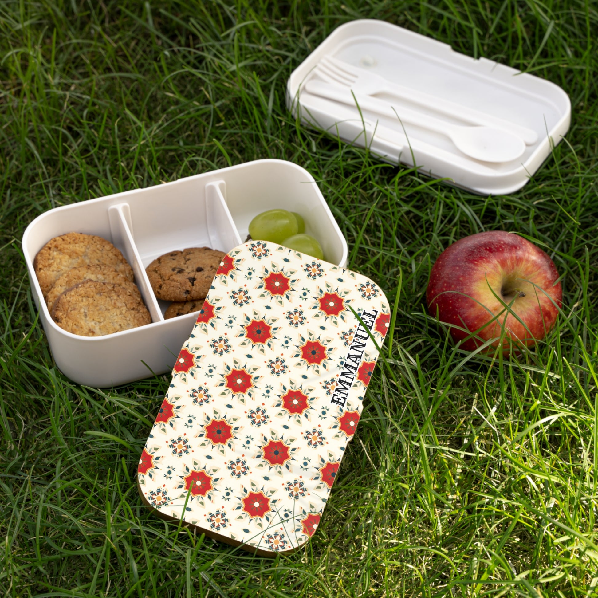 A floral-patterned, BPA-free lunchbox with three compartments—two with cookies, one with green grapes—sits on grass. The matching lid and a white utensil set are nearby. An apple rests beside this inviting picnic scene.
