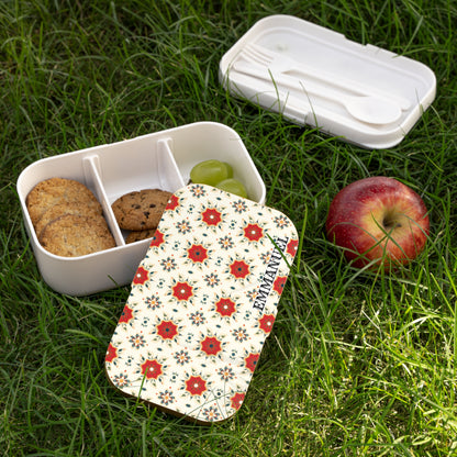 A floral-patterned, BPA-free lunchbox with three compartments—two with cookies, one with green grapes—sits on grass. The matching lid and a white utensil set are nearby. An apple rests beside this inviting picnic scene.