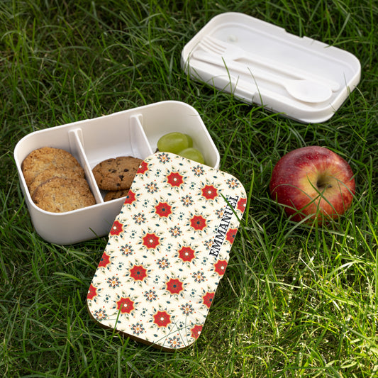 A floral-patterned, BPA-free lunchbox with three compartments—two with cookies, one with green grapes—sits on grass. The matching lid and a white utensil set are nearby. An apple rests beside this inviting picnic scene.