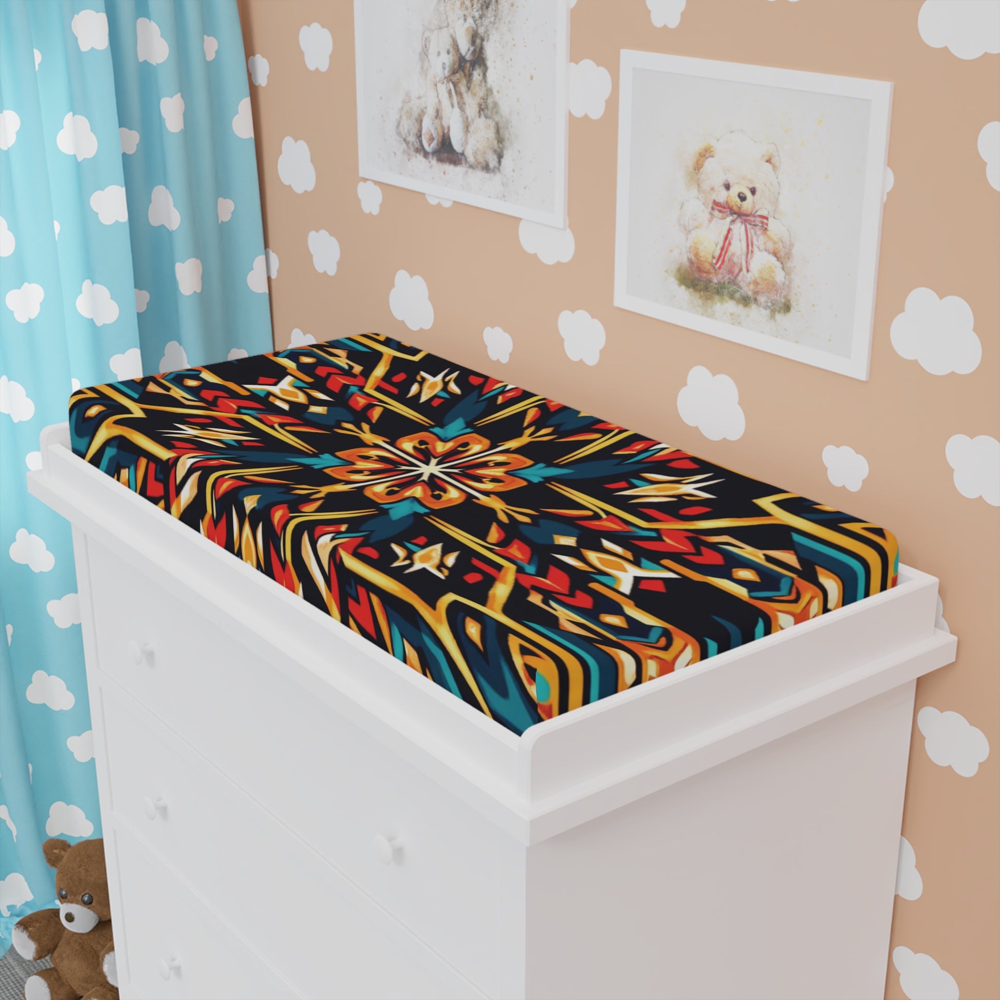 A colorful, African-inspired changing pad cover sits atop a white dresser in a child's room with tan walls, cloud decals, blue curtains with white clouds, framed teddy bear pictures on the wall, and a plush bear on the floor.