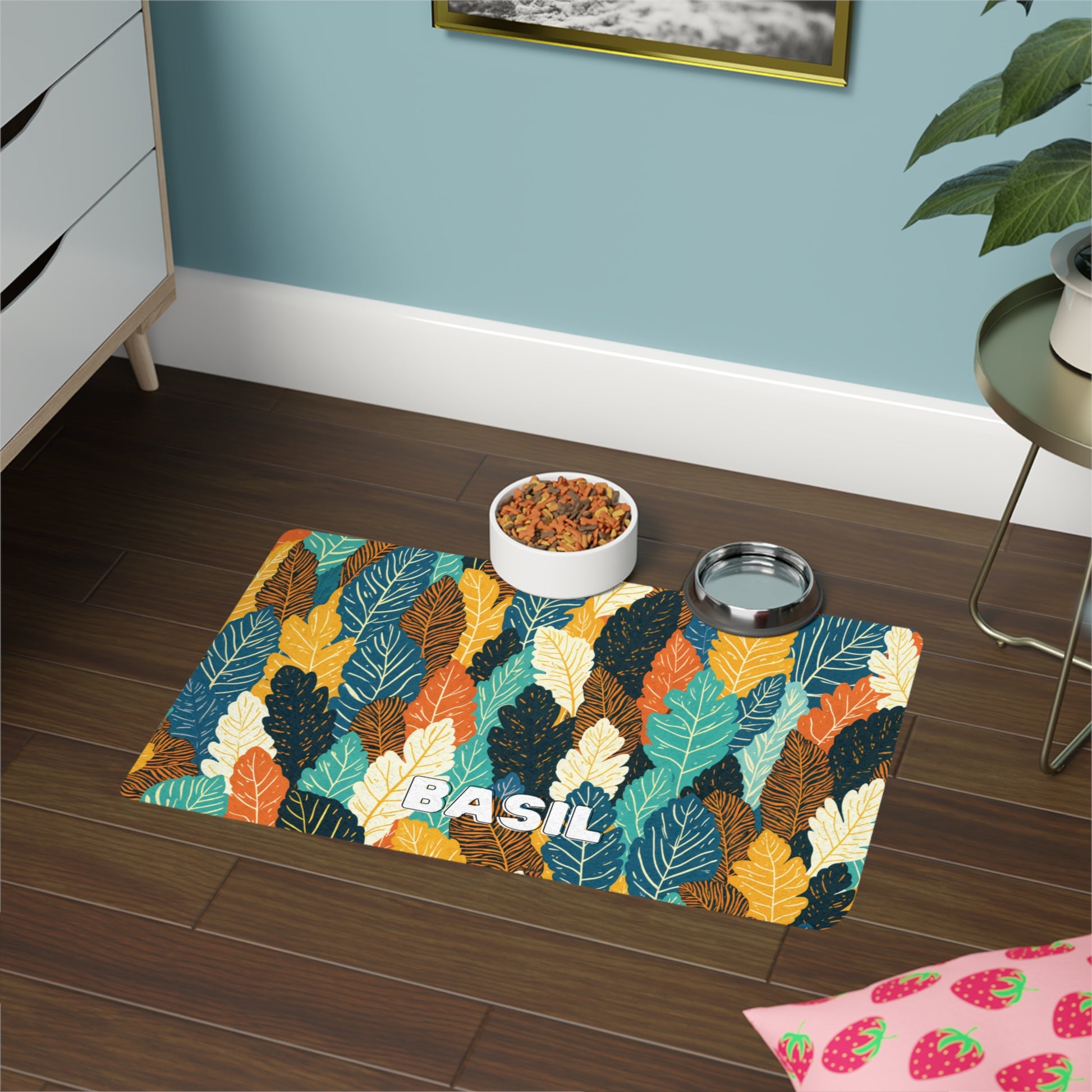 A colorful, African-inspired leaf-patterned mat labeled BASIL on a dark wooden floor holds a white pet food bowl with kibble and a metal water bowl. Nearby are a plant, a pink strawberry-patterned mat, and a dresser against a blue wall.