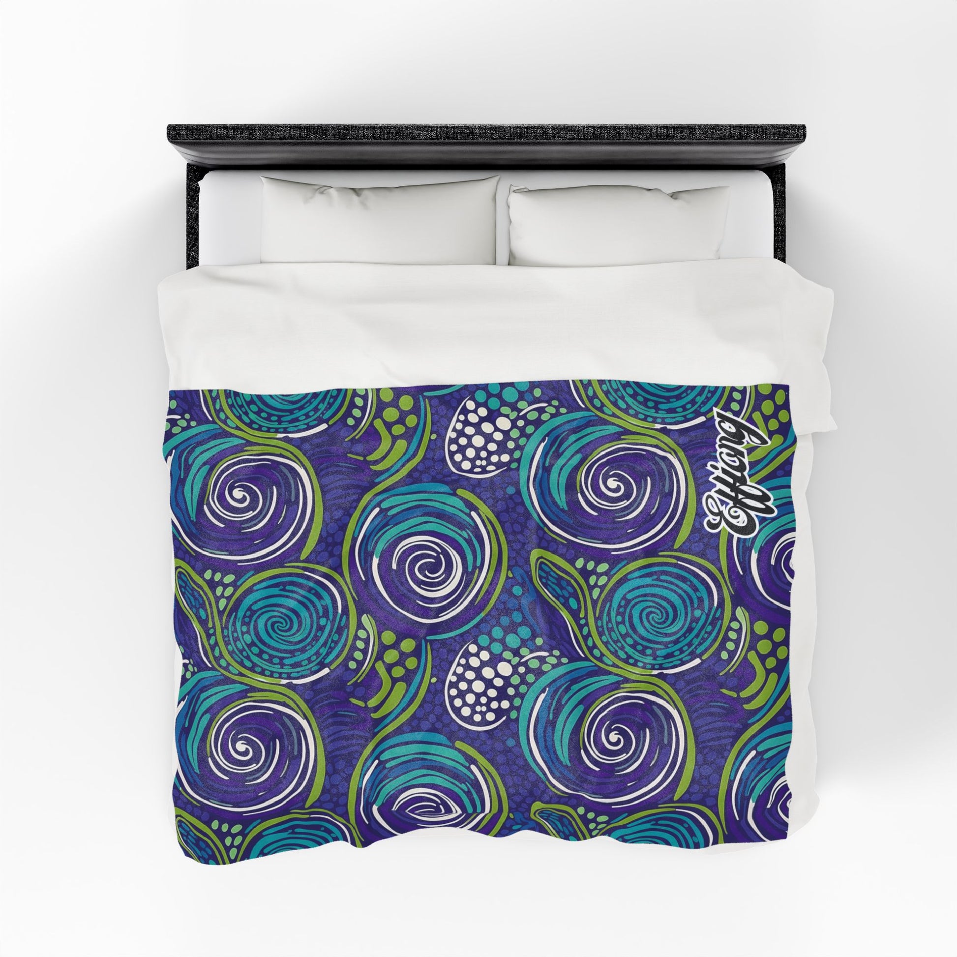 A bed with a white pillow and sheet set is topped by a personalized velveteen blanket featuring a vibrant, abstract swirl pattern in blue, purple, and green with white dotted accents and the Coastal logo. The photo is taken from an overhead view.