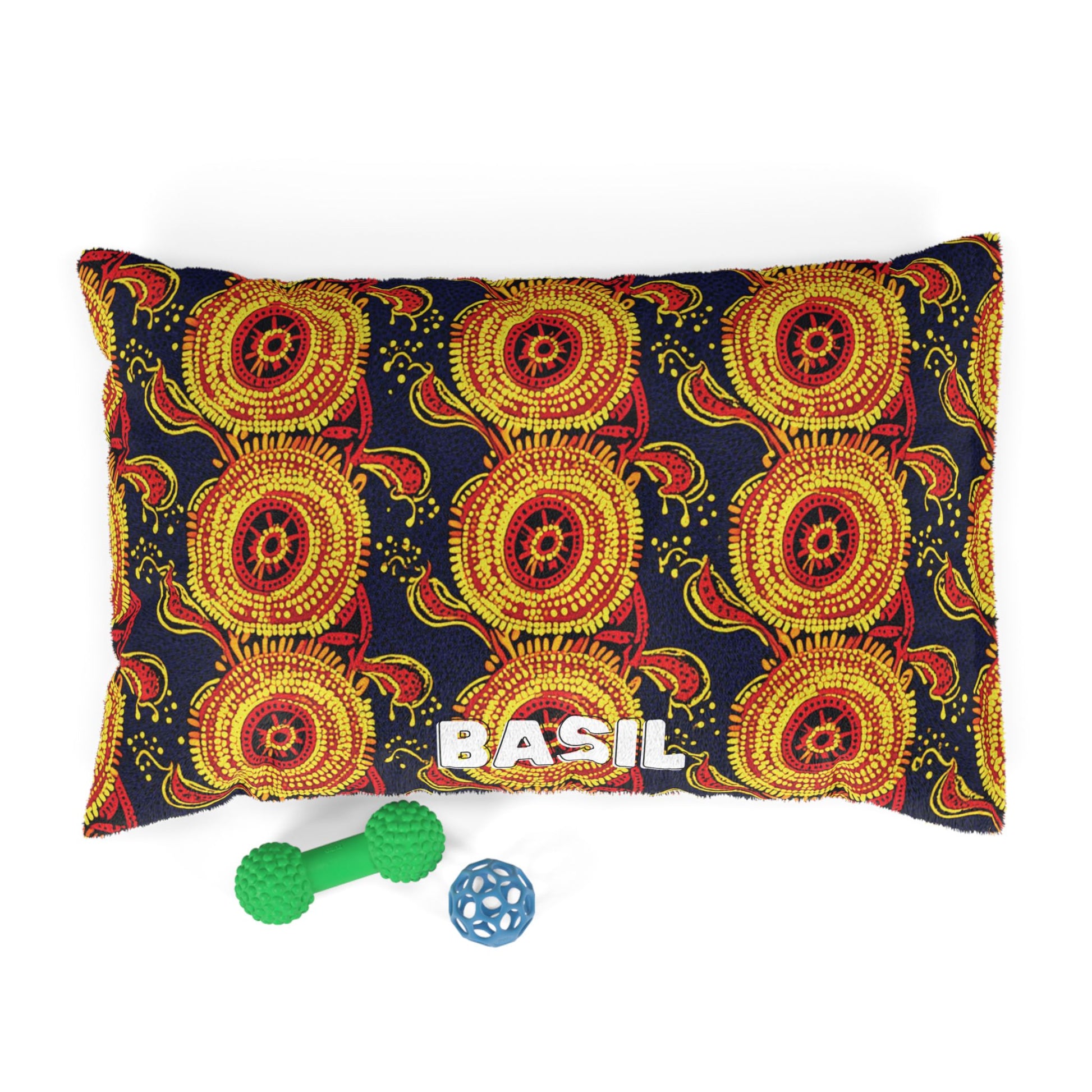 A rectangular pillow with a vibrant red, yellow, and orange African-inspired circular pattern on a black background. The name BASIL is printed in bold white letters at the bottom, with two green and blue dog chew toys in front.