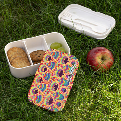 A BPA-free, African-inspired bento lunch box with a colorful, patterned lid and the word EXQUISITE sits on grass. The open box holds cookies, a grape, and an empty spot. Nearby are an apple and a white case with fork and spoon.
