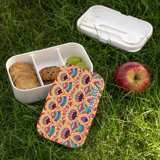 A BPA-free, African-inspired bento lunch box with a colorful, patterned lid and the word EXQUISITE sits on grass. The open box holds cookies, a grape, and an empty spot. Nearby are an apple and a white case with fork and spoon.