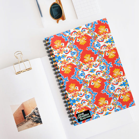 A vibrant journal with a red, yellow, and blue floral cover lies on an open book showing a wall and rocks. Nearby are tan sunglasses, a gold binder clip, and a small grid notepad arranged neatly on a white desk.