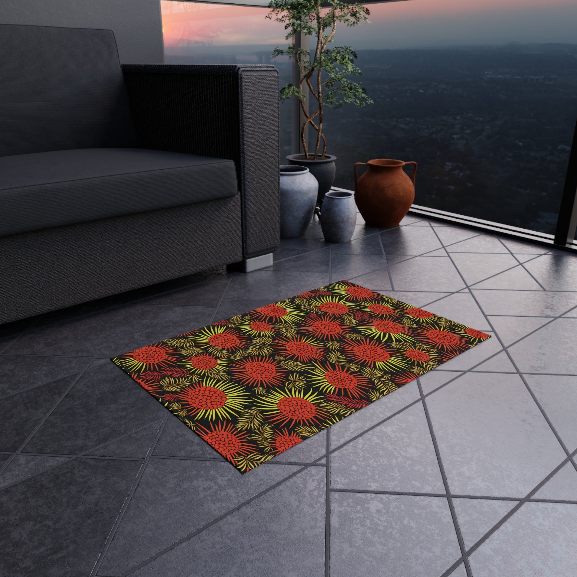 A small rectangular rug with a bold, African-inspired pattern of red and yellow spiky flowers lies on a tiled balcony floor. Nearby are an outdoor sofa, three ceramic pots, and a potted plant, with a cityscape visible at sunset.