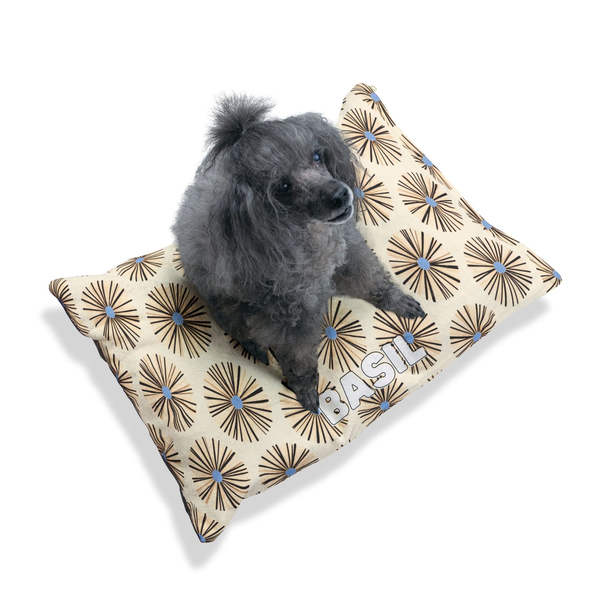 A small gray poodle sits on a beige rectangular pet bed with an African-inspired retro starburst pattern in brown, blue, and black. The word BASIL is printed in large white block letters on one corner. The background is plain white.