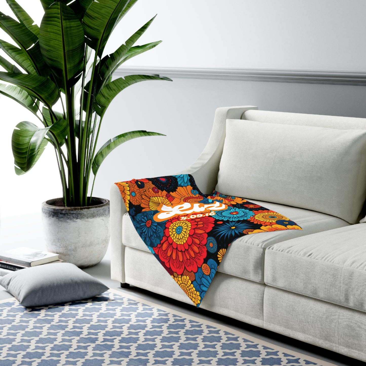 A white sofa with a colorful floral blanket, reminiscent of African-inspired decor, draped over the armrest sits next to a large potted plant. The bold blanket features red, yellow, and blue flowers with “Let’s” in white script. A patterned blue and white rug completes the look.