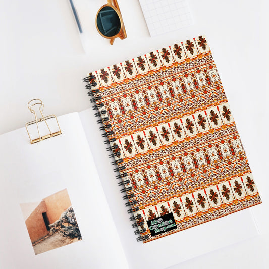 A premium writing notebook with a floral and geometric cover, inspired by African design, lies on an open book showing a building photo. Nearby are brown sunglasses, a gold binder clip, and graph paper on a clean white surface.