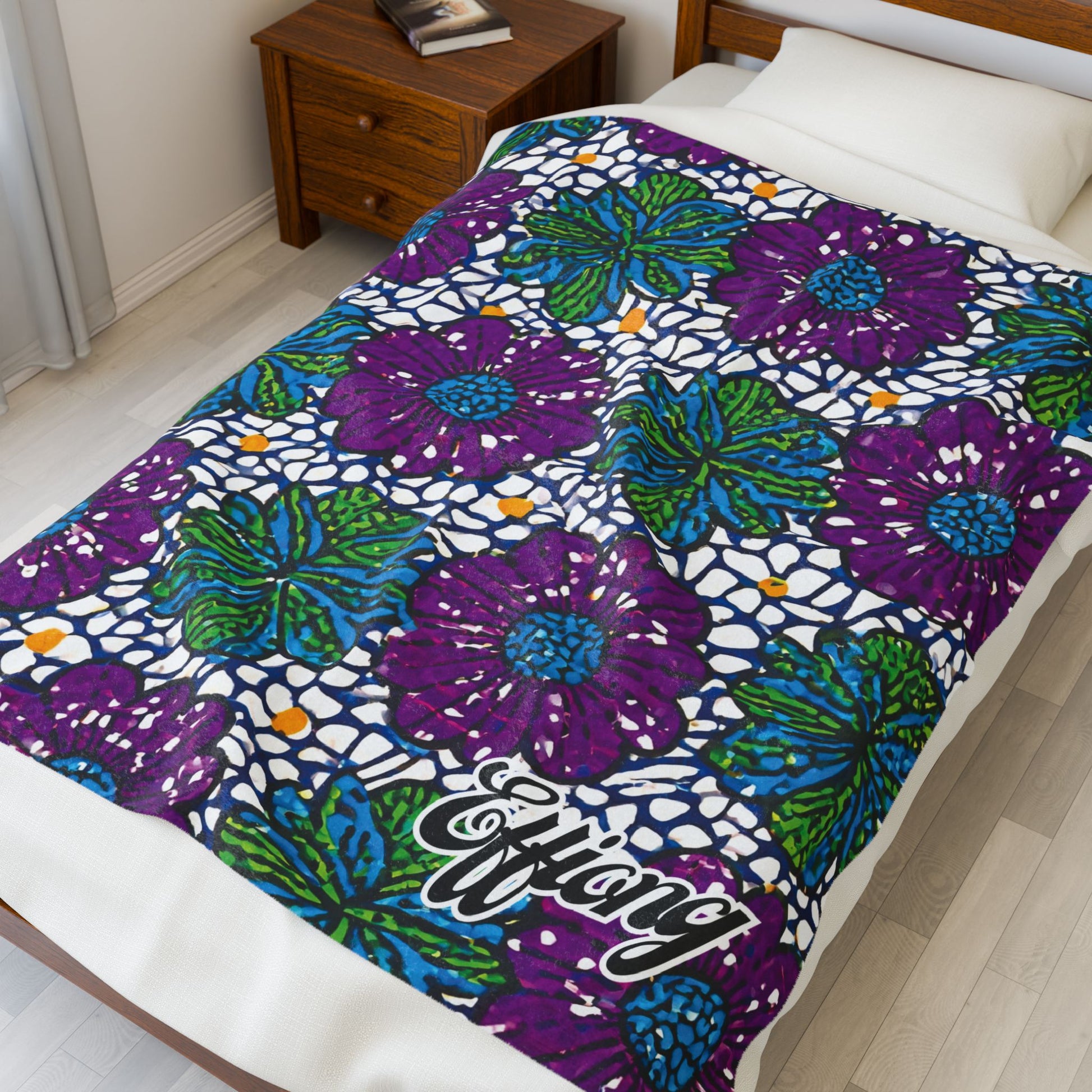 A bed is covered with an African-inspired personalized blanket featuring large purple and green flowers with blue and orange accents on white. “Efiang” is printed in script at the bottom. A wooden nightstand with books sits on a light wood floor.