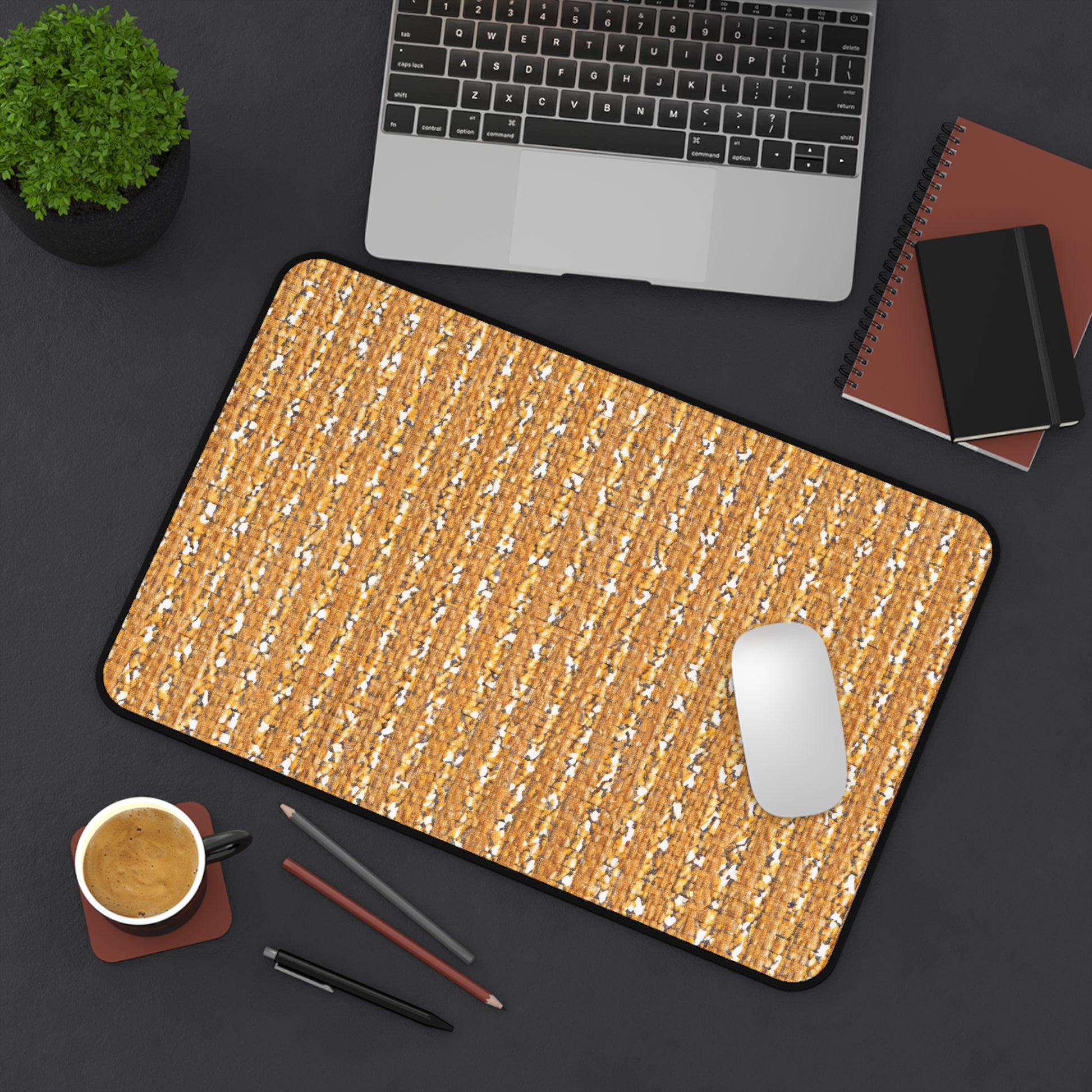 A tan and white ergonomic desk pad with a textured pattern sits on a dark desk, alongside a silver laptop, a white mouse, a brown coffee cup and saucer, two notebooks, two pencils, a pen, and a small green potted plant.