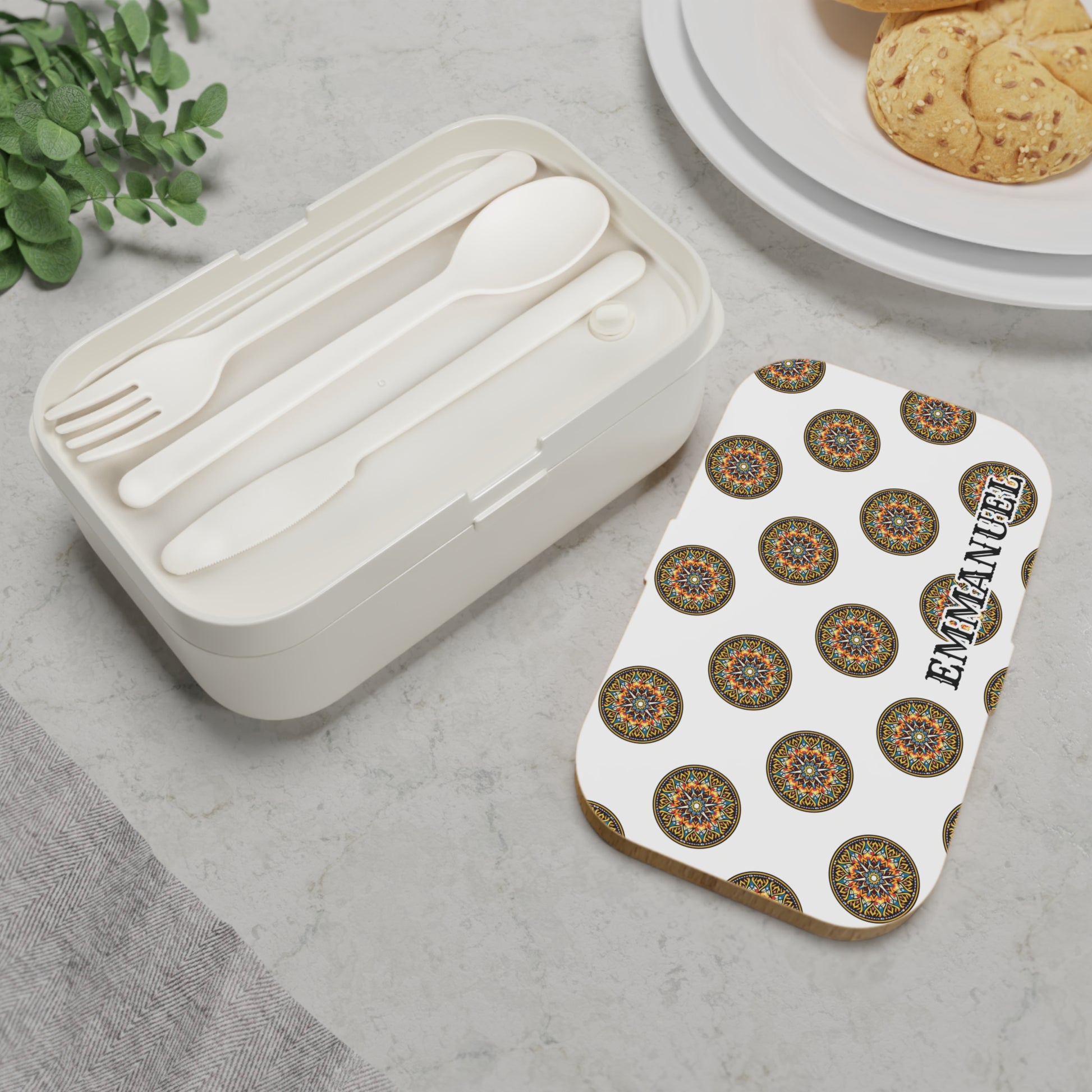 A personalized, BPA-free lunchbox with a colorful mandala lid and EMMANUEL in bold text sits on marble. This African-inspired bento lunch box includes a built-in compartment for utensils, with plates of cookies arranged nearby.