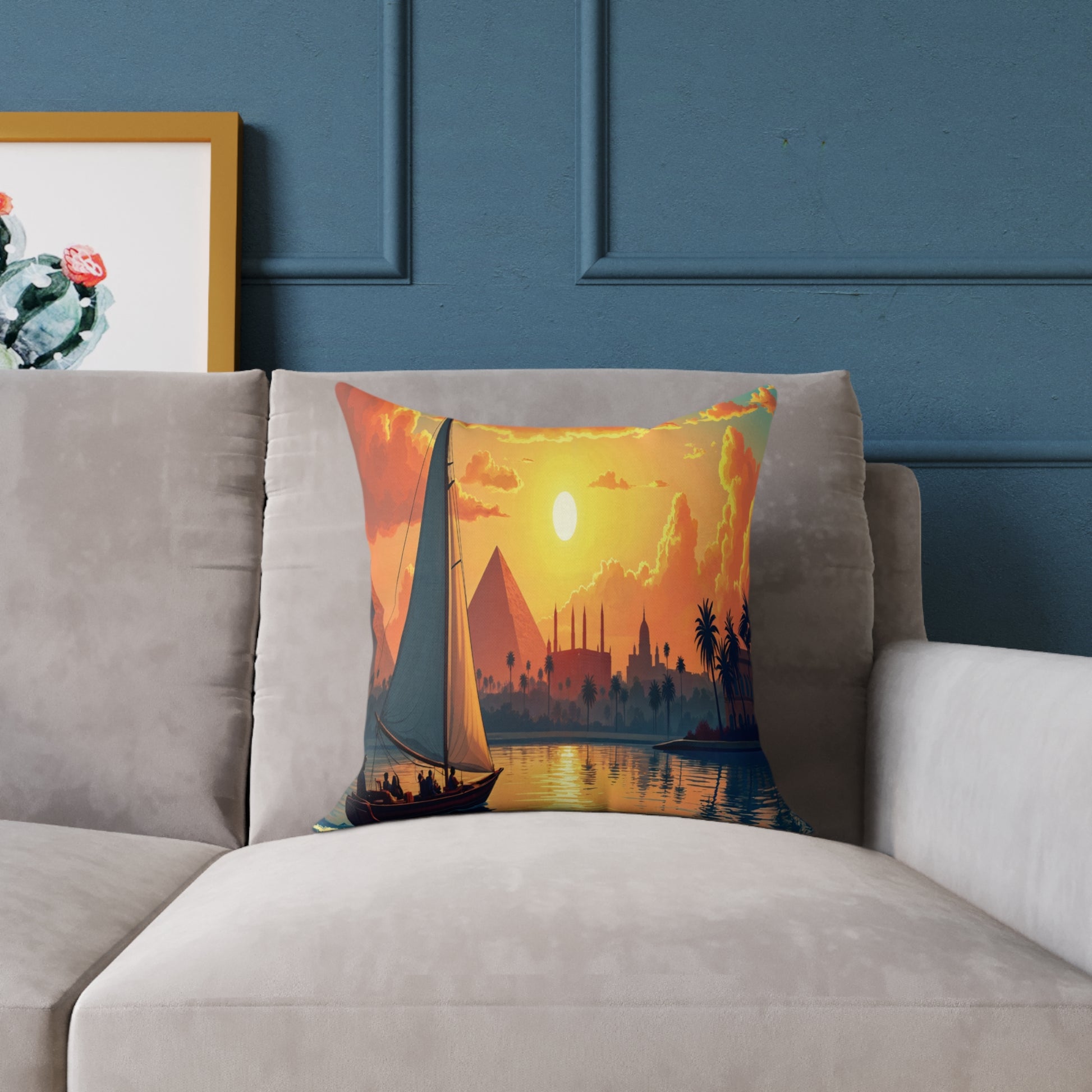 A vibrant throw pillow on a beige couch features a sailboat at sunset, with pyramids, palm trees, and mosque silhouettes. Blue paneled walls and framed cactus art add charm to this colorful home decor setting.
