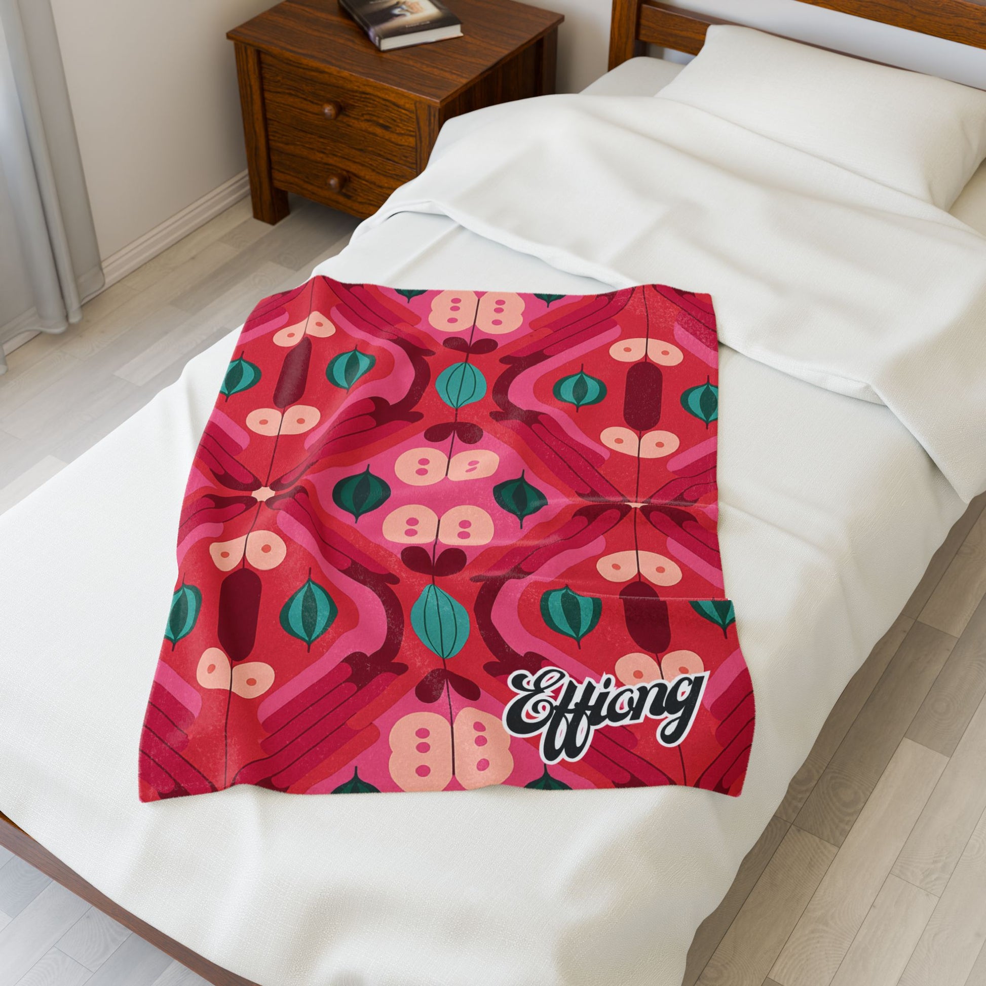 A colorful, African-inspired blanket with bold abstract patterns in red, pink, green, and teal lies on a neatly made white bed. The personalized velveteen blanket features Effiong in large white script. A wooden nightstand with books is beside the bed.