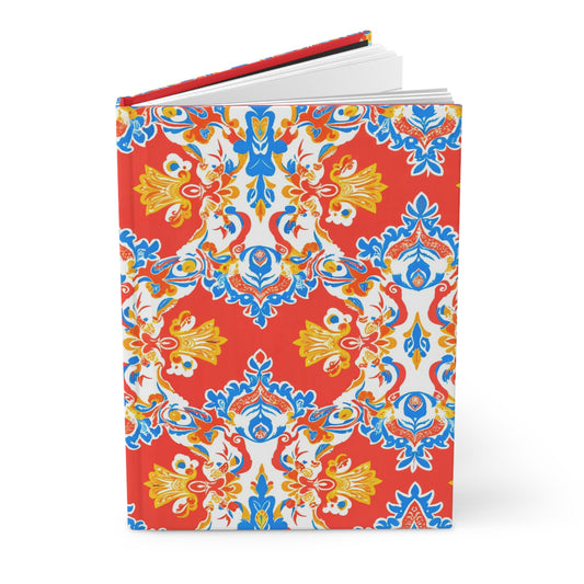 A hardcover journal with a vibrant cover featuring an ornate, symmetrical floral pattern in blue, yellow, and white on a bright red background. The African-inspired journal stands upright with pages slightly open, revealing a red inner cover.