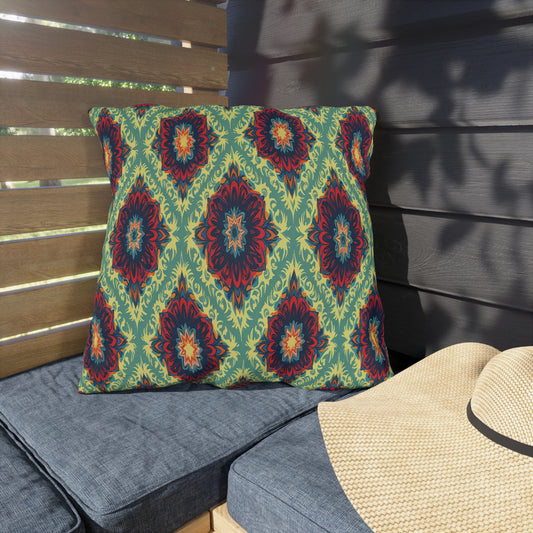 A square cushion with an African-inspired geometric and floral pattern in green, blue, orange, and red rests on a gray outdoor bench. Beside it is a straw hat. Wooden slats and dark siding create a shaded, cozy patio setting.