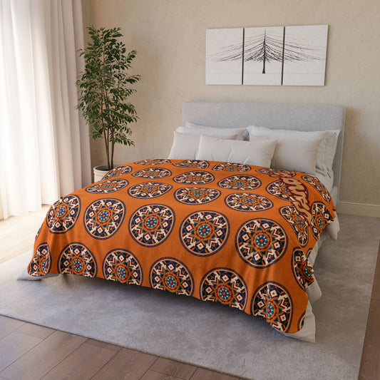 A neatly made bed with a grey headboard is topped with an orange, African-inspired blanket featuring large, colorful mandala patterns. The room has a light grey rug, wooden floor, a green potted plant, and abstract black-and-white wall art above the bed.