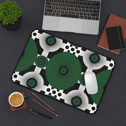 A large neoprene desk mat with a bold black, white, and green geometric pattern sits on a dark desk. Nearby are a laptop, notebooks, a smartphone, pens, coffee, and a potted plant. A white mouse rests on this colorful workspace accessory.