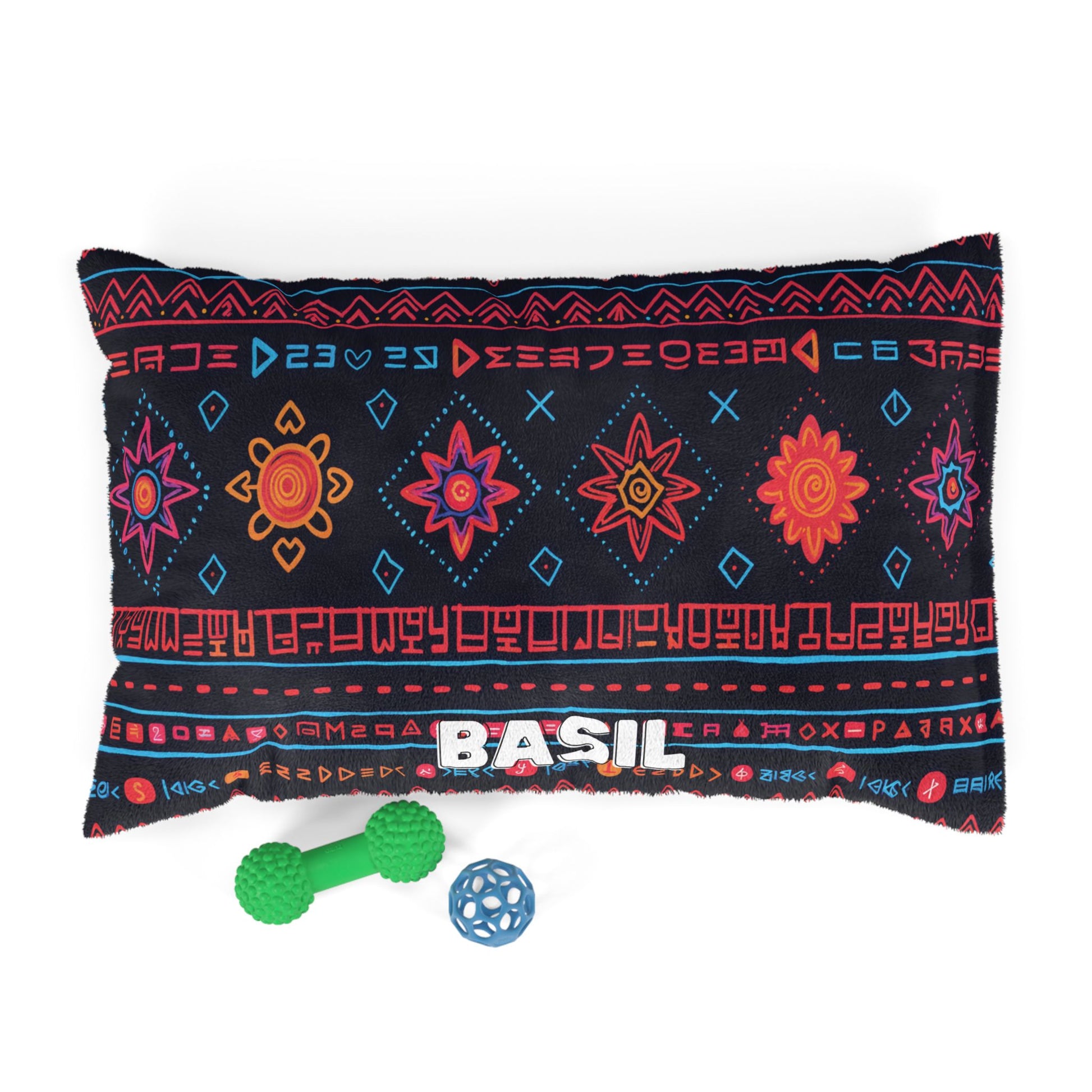 A black rectangular pillow showcases colorful geometric and floral patterns, with the name BASIL in bold white letters—perfect for personalized pet beds. Below are a green bone-shaped toy and a small blue ball with holes, all set against a white background.