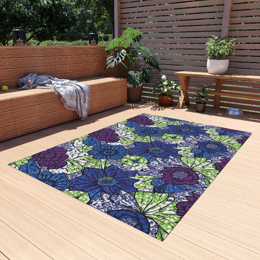 A colorful patio rug with large blue, green, and purple floral patterns lies on a wooden deck. Nearby are a wicker sofa with a gray blanket, leafy potted plants, and a wooden table. Trees and greenery are visible in the background.