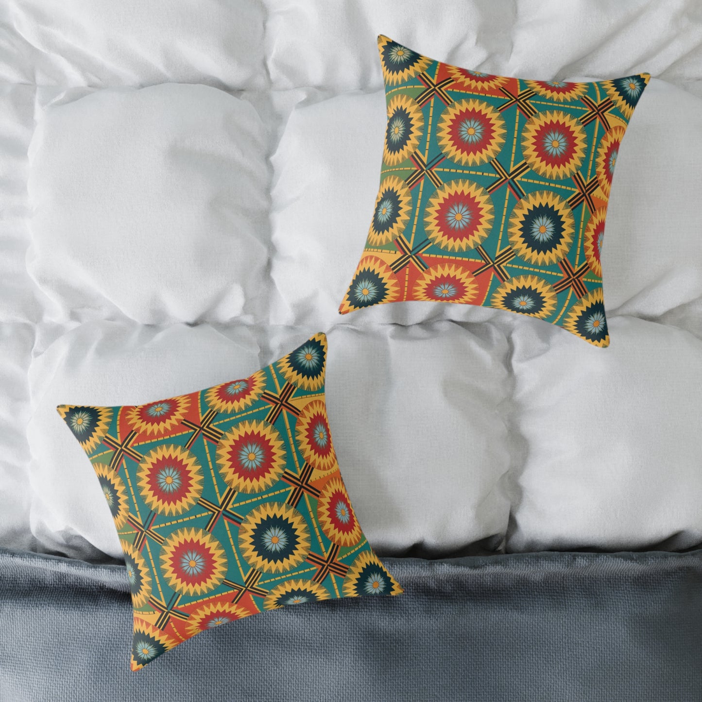 Two vibrant throw pillows with a colorful geometric starburst pattern in yellow, red, teal, and black rest on a white, tufted comforter. Part of a gray, textured blanket is visible at the bottom edge of this colorful home decor scene.