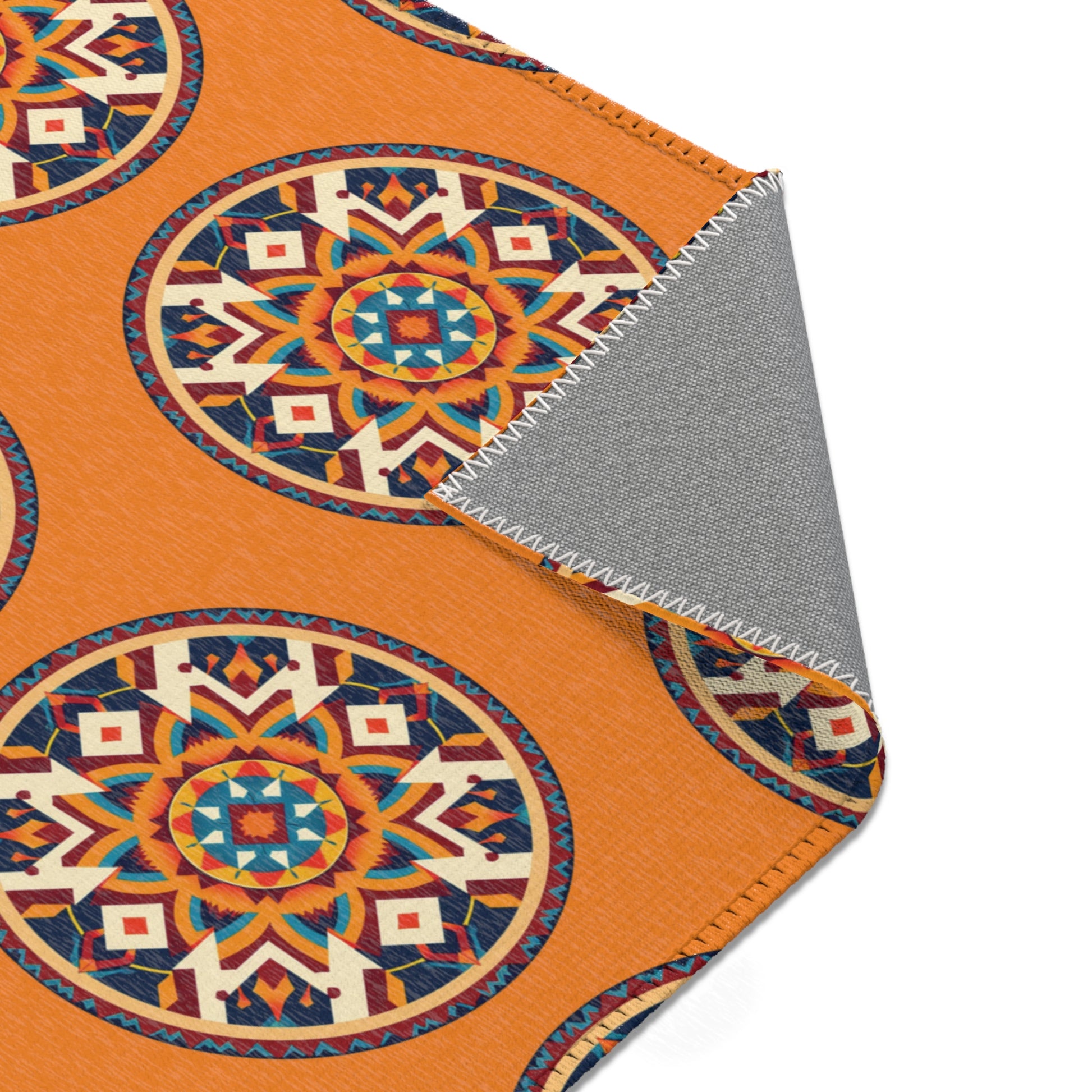 A corner of an orange, African-inspired blanket with large, circular, geometric Aztec-style patterns in blue, red, and cream. The gray underside is visible at the folded corner, showing white zigzag stitching along the edge.