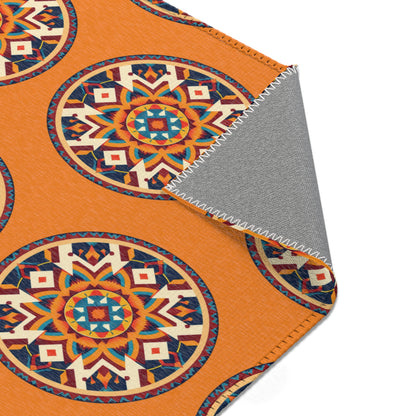 A corner of an orange, African-inspired blanket with large, circular, geometric Aztec-style patterns in blue, red, and cream. The gray underside is visible at the folded corner, showing white zigzag stitching along the edge.