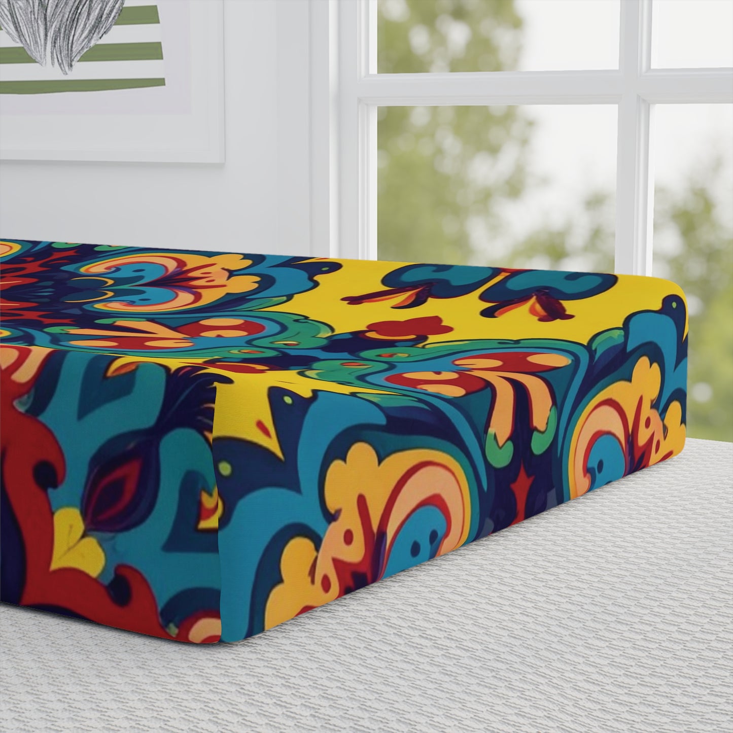 A vibrant mattress with a bold, colorful mandala pattern in yellow, blue, red, and orange sits on a bed by a sunlit window. Inspired by African-inspired designs, it pairs beautifully with custom changing pad covers for a unique nursery touch.