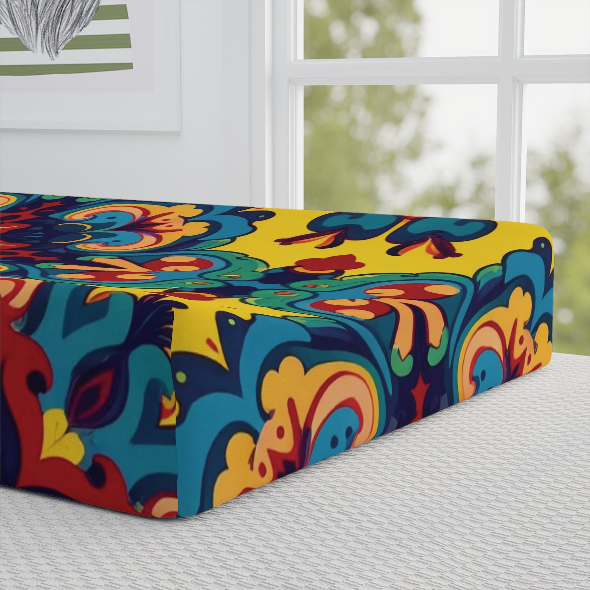 A vibrant mattress with a bold, colorful mandala pattern in yellow, blue, red, and orange sits on a bed by a sunlit window. Inspired by African-inspired designs, it pairs beautifully with custom changing pad covers for a unique nursery touch.