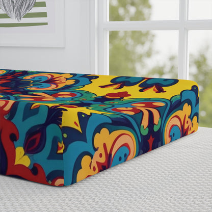 A vibrant mattress with a bold, colorful mandala pattern in yellow, blue, red, and orange sits on a bed by a sunlit window. Inspired by African-inspired designs, it pairs beautifully with custom changing pad covers for a unique nursery touch.