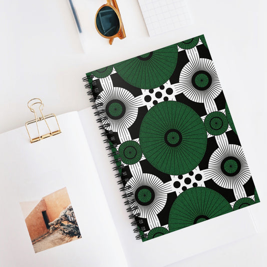A premium writing notebook with a bold green, black, and white geometric pattern lies on a white desk. Nearby are an open book with a photo, brown sunglasses, a gold binder clip, and a small checkered notepad.