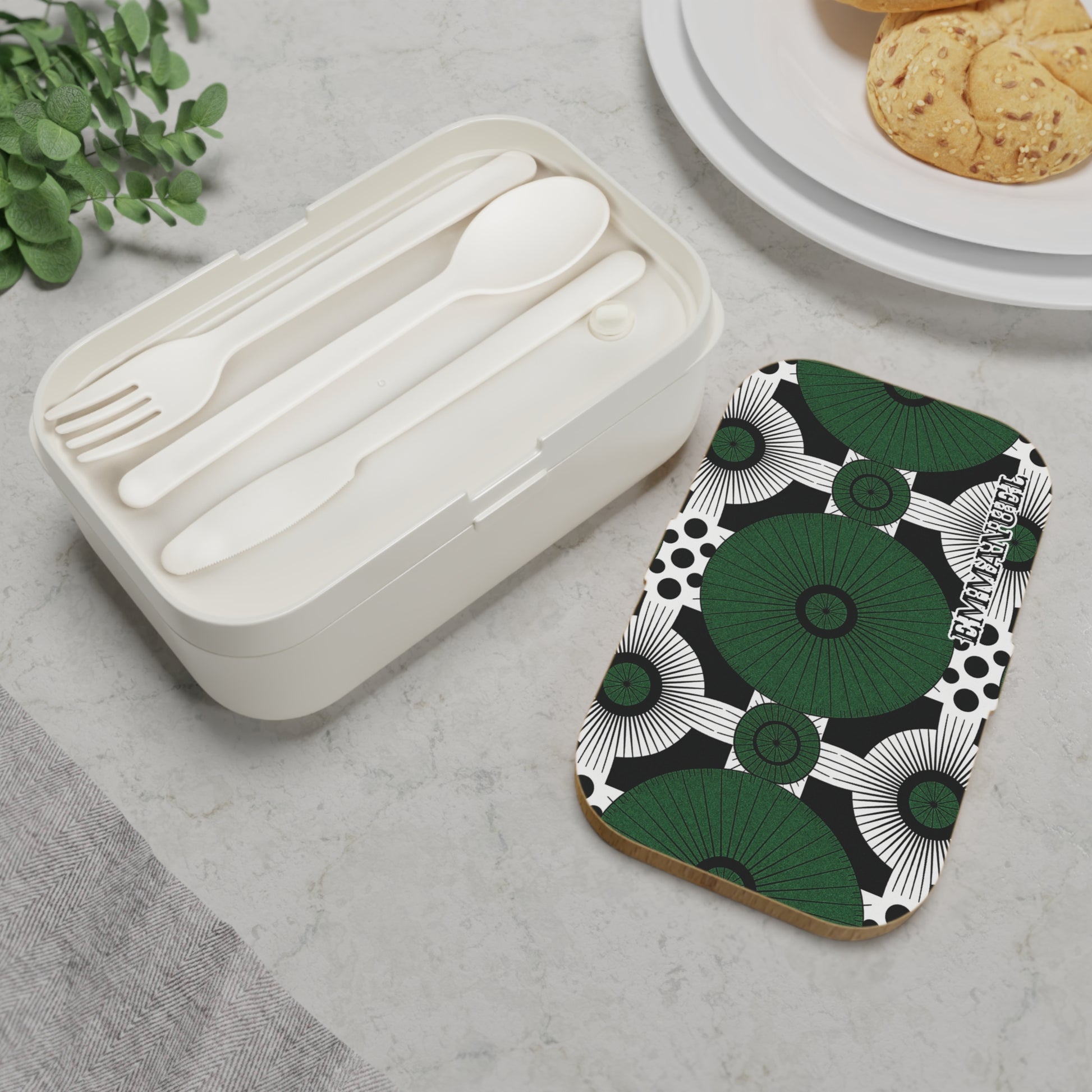 A white personalized bento lunch box with a matching spoon, fork, and knife sits on a marble surface next to a rectangular item featuring a bold green, black, and white abstract pattern. Part of a plate with cookies and greenery are also visible.
