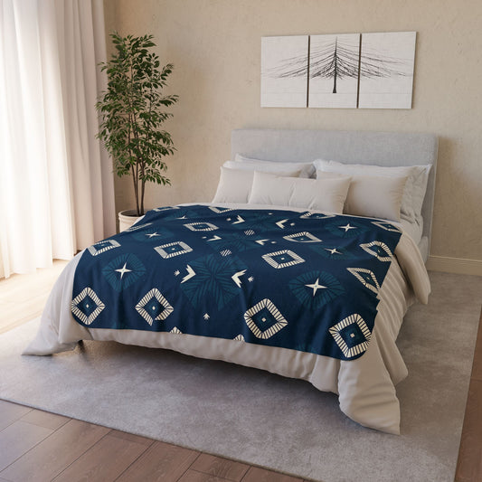 A neatly made bed with a navy blue quilt featuring geometric white patterns. Accented with cozy home decor, the room includes several white pillows, a light gray headboard, and a potted plant beside the bed, plus minimalist black-and-white tree artwork above.