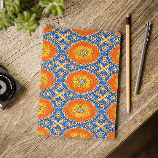 A vibrant African-inspired journal with bold orange, blue, and yellow geometric patterns lies on a wooden table beside a pencil, silver pen, camera, and a green potted plant partially visible in the top left corner.