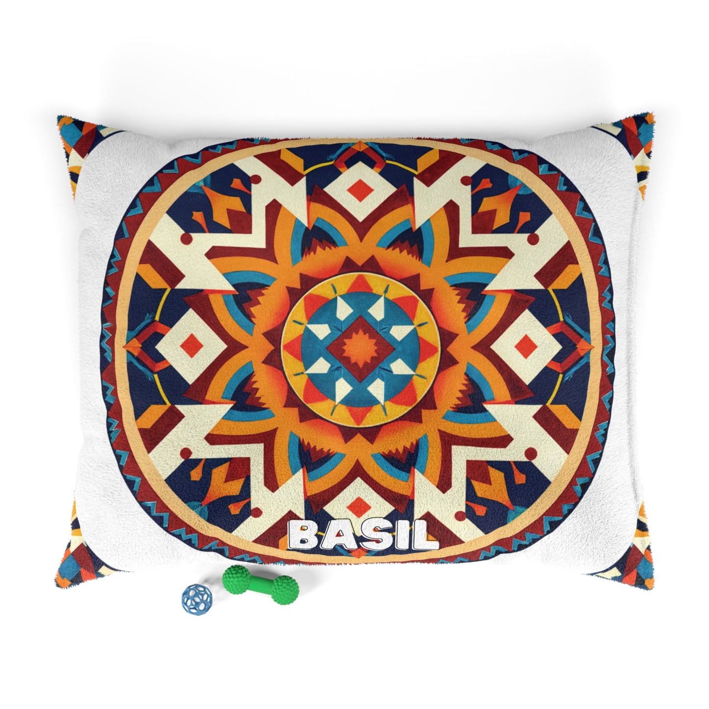 A decorative pillow with a vibrant geometric mandala design in orange, blue, cream, and red. The word BASIL is printed in bold white letters at the bottom center, offering a touch of luxurious comfort. Small green and blue dog toys rest nearby on a white background.