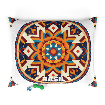 A decorative pillow with a vibrant geometric mandala design in orange, blue, cream, and red. The word BASIL is printed in bold white letters at the bottom center, offering a touch of luxurious comfort. Small green and blue dog toys rest nearby on a white background.