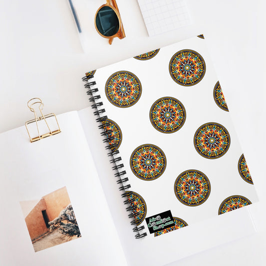 A spiral-bound notebook with a white cover features vibrant, colorful mandala patterns inspired by African designs. It rests on a white desk beside an open book with a photo, a gold binder clip, sunglasses, and a small grid notepad.