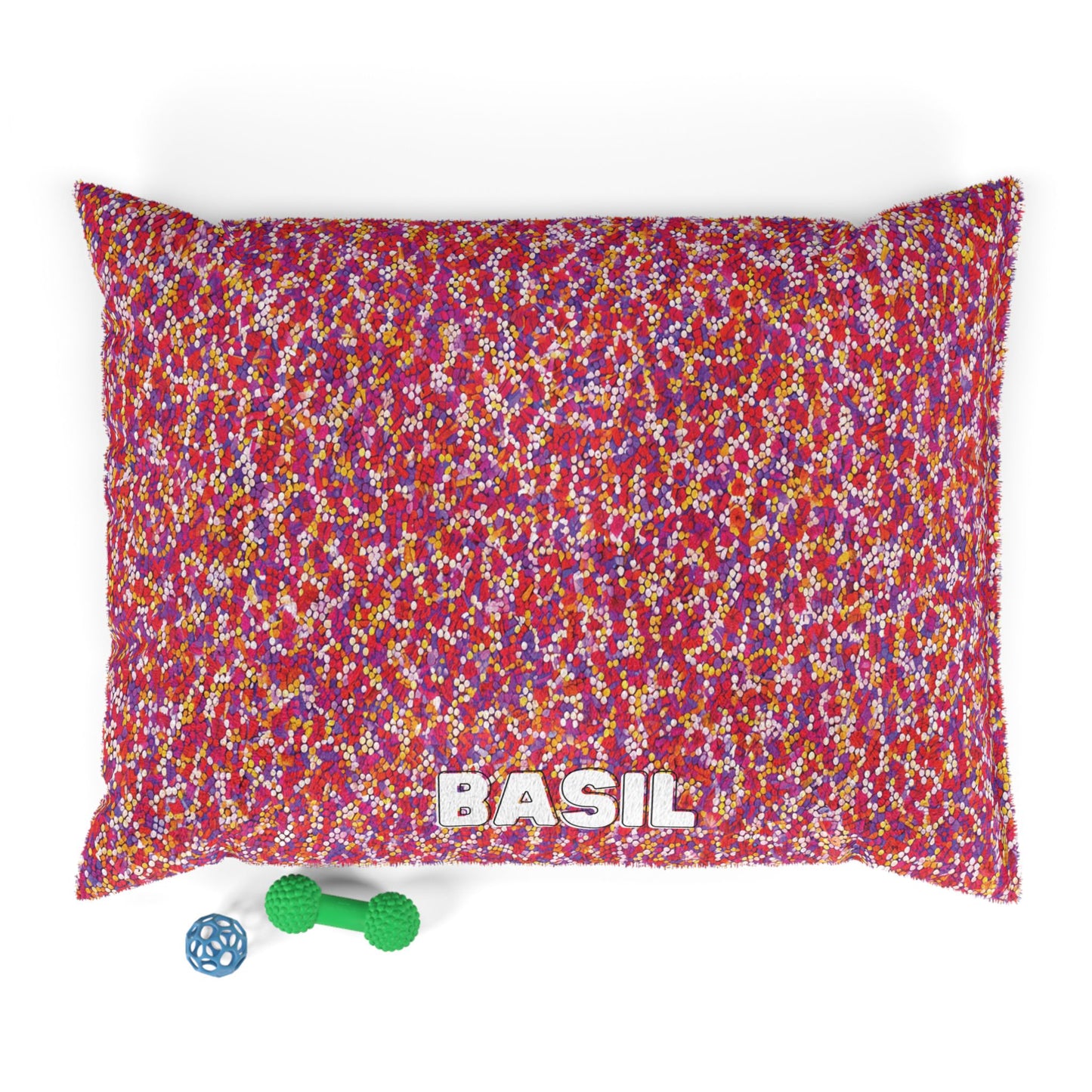 A rectangular pillow with a colorful, speckled pattern in red, pink, yellow, and white, labeled BASIL in white letters at the bottom. Enjoy luxurious comfort for your pet—plus two dog toys: a green dumbbell-shaped toy and a blue ball with holes.