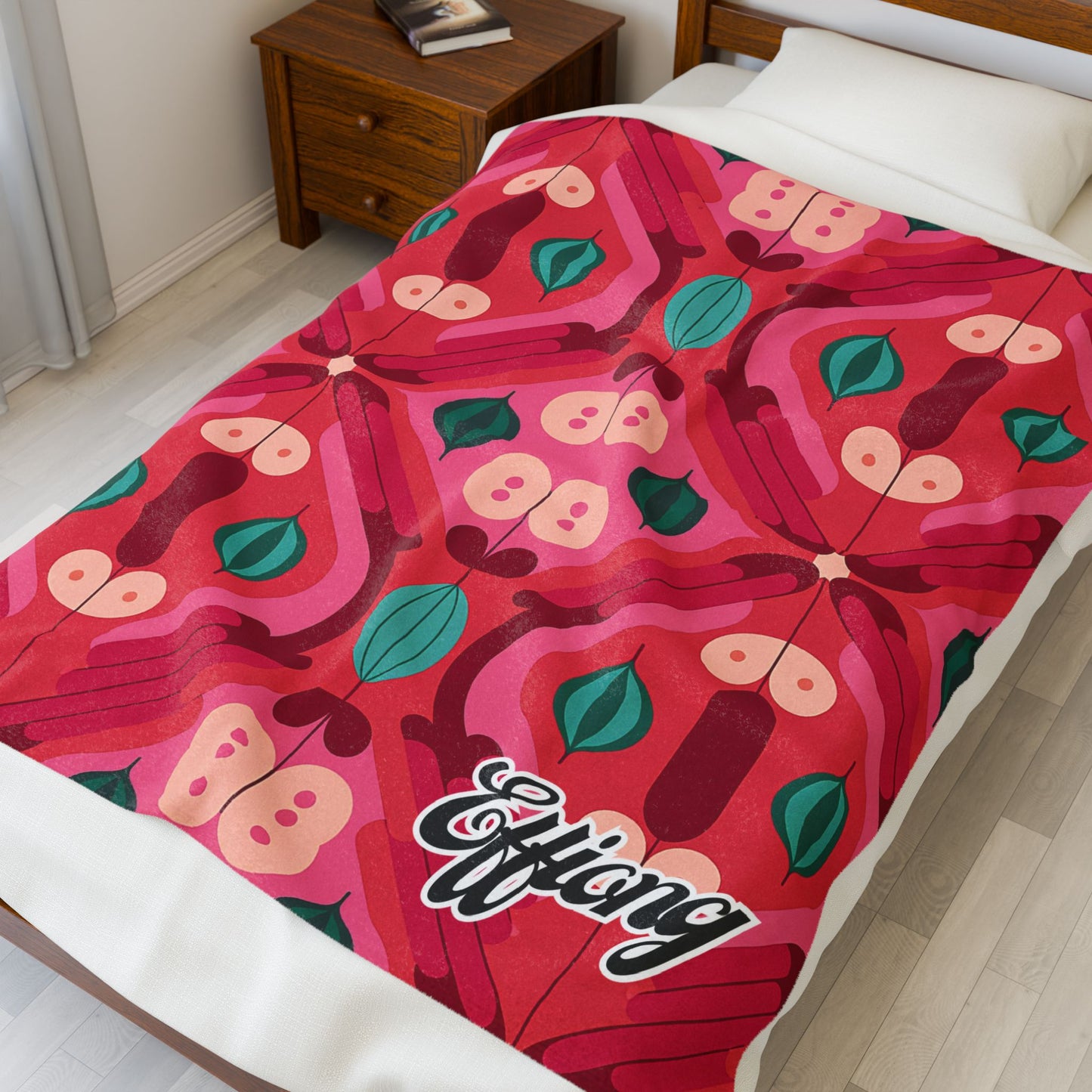 A bed is covered with a vibrant African-inspired blanket featuring a symmetrical butterfly and leaf pattern in red, pink, and green. The personalized velveteen blanket has Efflang in stylized script at the corner. A wooden nightstand sits beside the bed.
