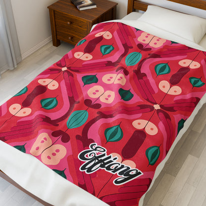 A bed is covered with a vibrant African-inspired blanket featuring a symmetrical butterfly and leaf pattern in red, pink, and green. The personalized velveteen blanket has Efflang in stylized script at the corner. A wooden nightstand sits beside the bed.