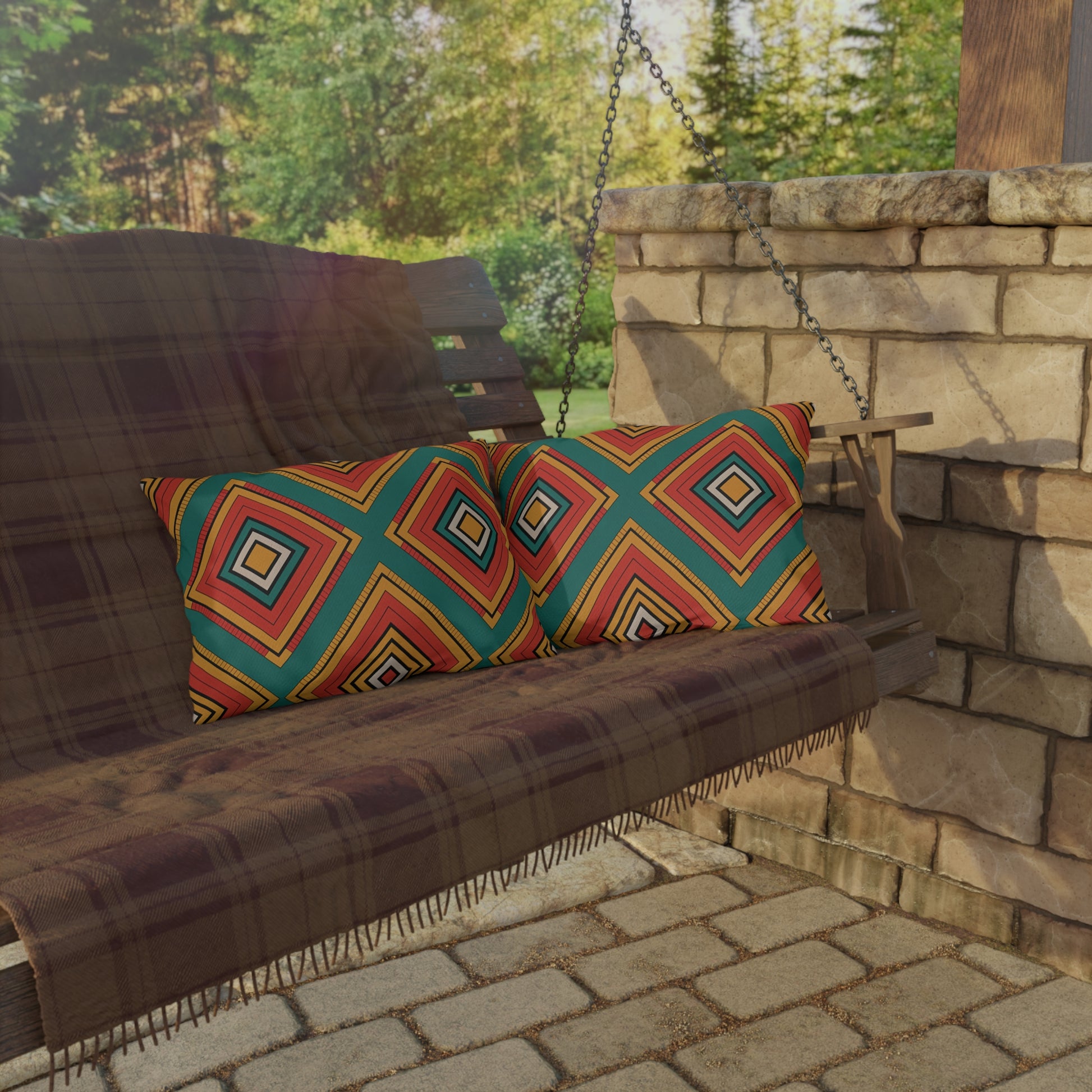 A wooden porch swing with a plaid blanket and outdoor pillows featuring colorful, geometric patterns sits on a stone patio by a stone wall, hanging from metal chains. Green trees and sunlight create a relaxing backdrop for this cozy spot.
