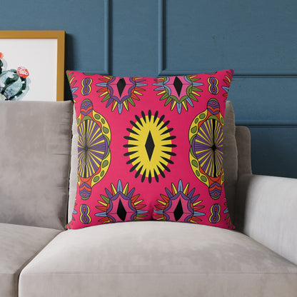 A square African-inspired pillow with a bold, colorful geometric and abstract pattern in yellow, black, orange, and purple on pink rests on a light gray sofa against a blue paneled wall. Perfect as a vibrant home decor accent. Framed art peeks in from the left.