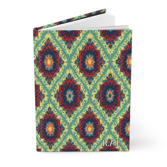 A hardcover monogram journal with an ornate, symmetrical floral pattern in red, blue, and yellow on a green background. The book is partly open, and the initials R.A.E. are printed in white at the bottom right corner of the front cover.