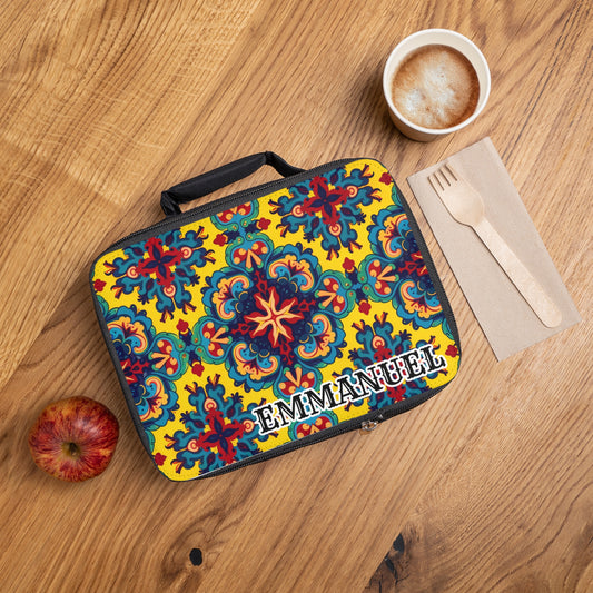 A vibrant personalized lunch bag with colorful floral and geometric patterns sits on a wooden table. The name “EMMANUEL” is printed in white. Beside this African-inspired lunch tote are a red apple, cappuccino, and wooden cutlery on a napkin.