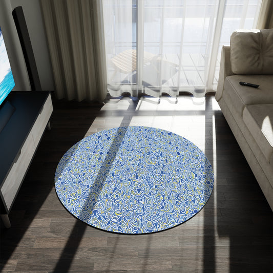 A round, African-inspired rug with a busy blue, white, and yellow abstract pattern lies on a dark wood floor between a light sofa and TV stand. Sunlight streams through sheer curtains, brightening the room as a remote rests on the sofa.